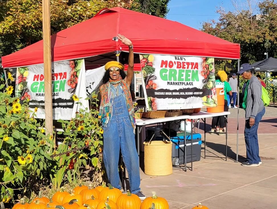 Beverly Grant waving beneath the red Mo'Betta Green canopy at the market, sunflowers and pumpkins in the foreground.